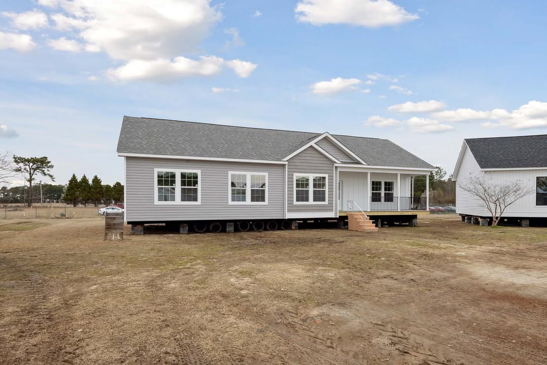 A gray manufactured home sits on a barren lot under a partly cloudy sky. It features white trim, multiple windows, and a small front porch with steps.