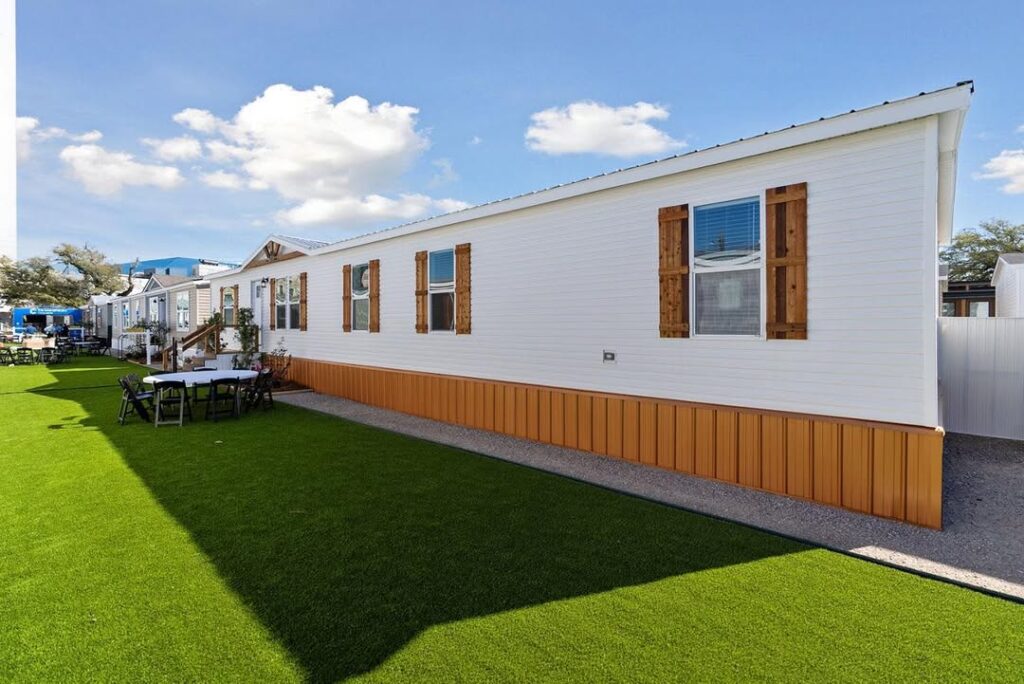 A white, single-story mobile home with wooden shutters sits on neat green artificial grass under a blue sky with scattered clouds, conveying a serene atmosphere.