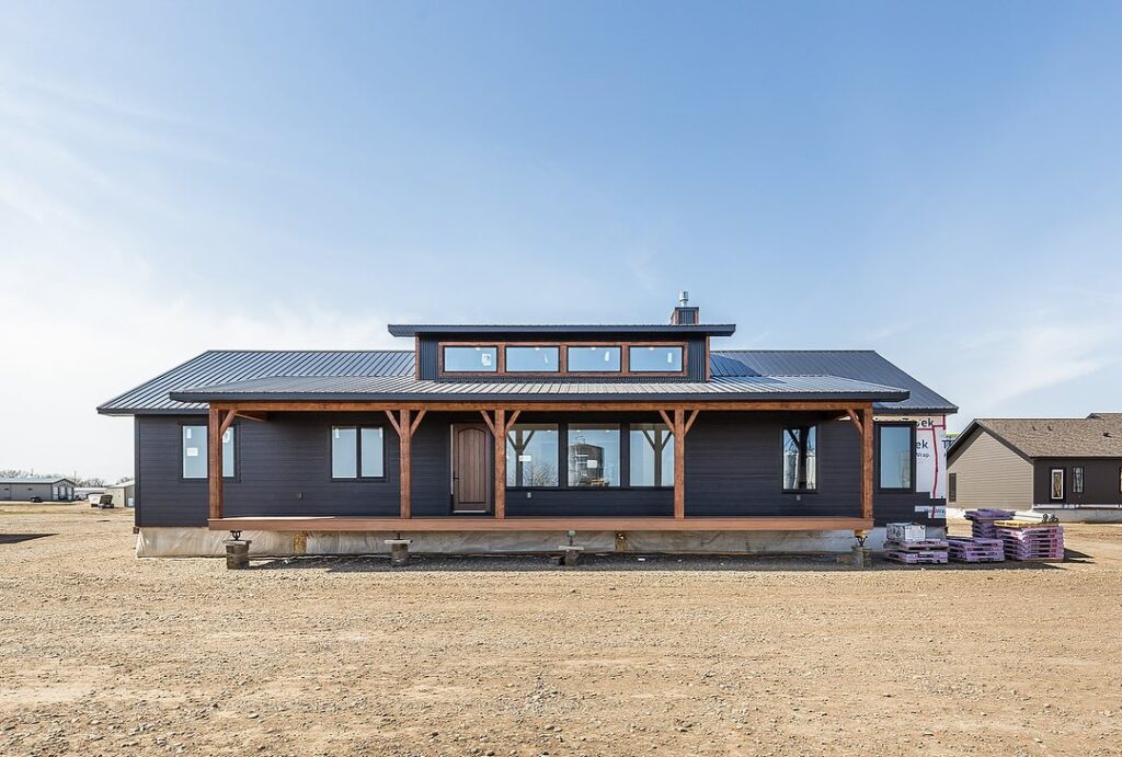 A modern, single-story black house with large windows and wooden beams is surrounded by an open dirt area under a clear blue sky, exuding a calm, rustic ambiance.