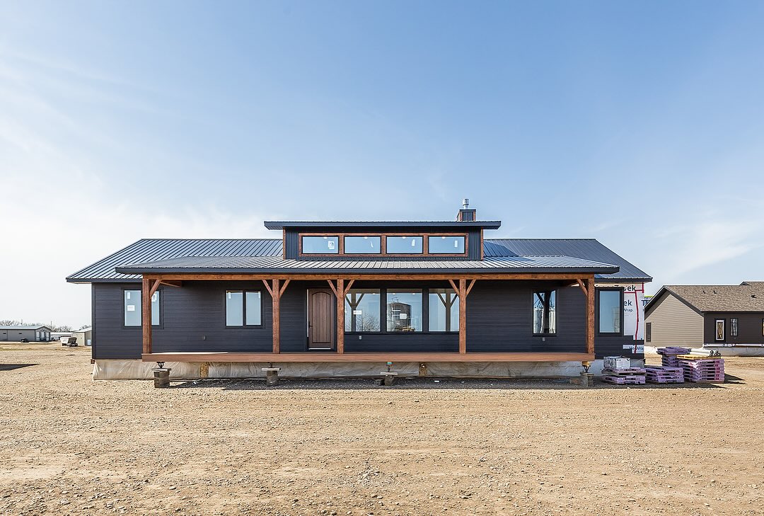 A modern, single-story black house with large windows and wooden beams is surrounded by an open dirt area under a clear blue sky, exuding a calm, rustic ambiance.