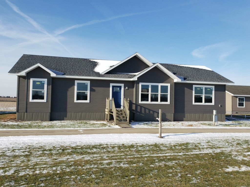 A single-story gray house with a dark roof, front steps, and several windows. Snow covers parts of the lawn. The sky is clear and blue.