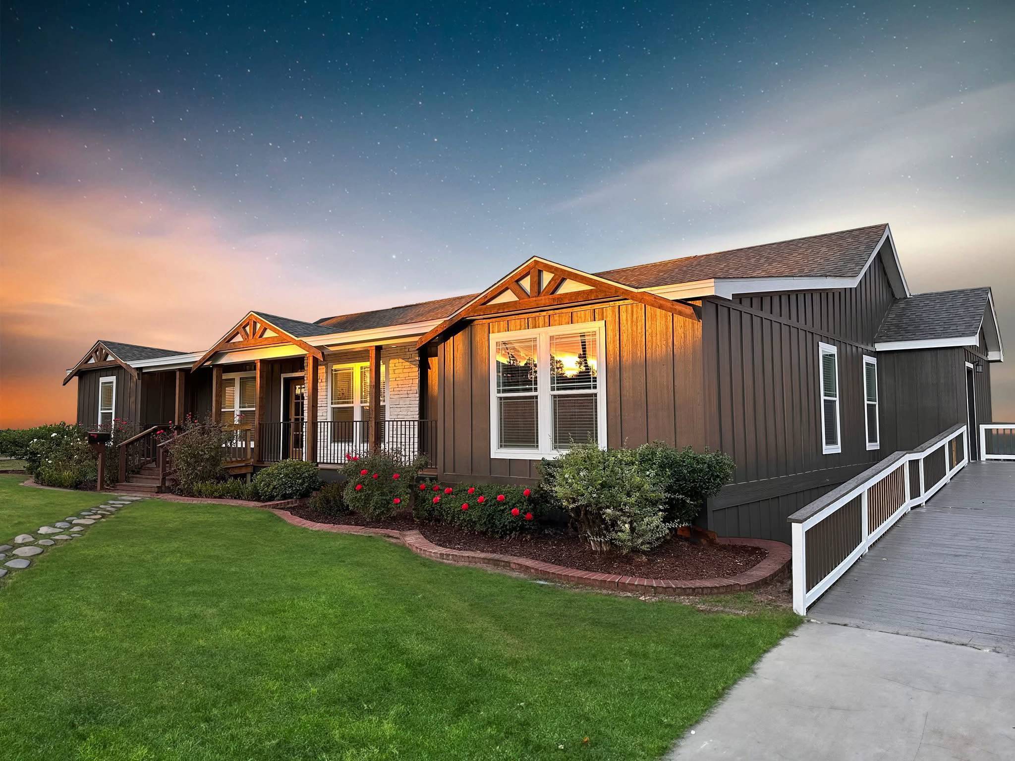 Single-story house at sunset with illuminated windows, surrounded by a manicured lawn and flowerbed. Clear evening sky with a tranquil ambience.