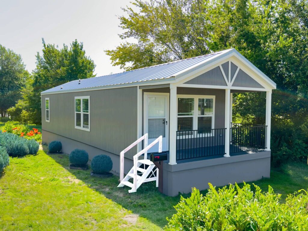 Small grey mobile home with white trim and a metal roof, set in a lush green yard. A small porch and steps lead to the front door, conveying a cozy feel.