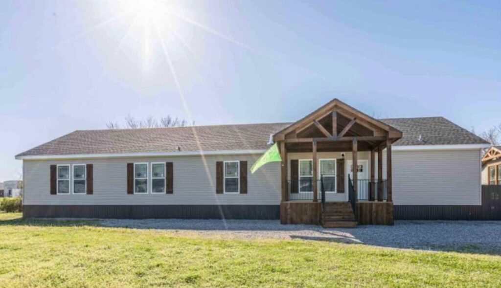 A light gray, single-story house with a brown gabled porch sits under a bright sun. It has several windows and is surrounded by a green lawn.