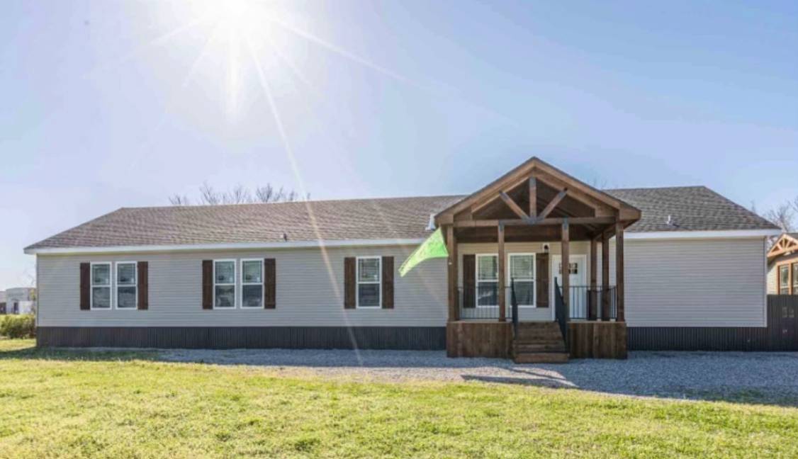 A light gray, single-story house with a brown gabled porch sits under a bright sun. It has several windows and is surrounded by a green lawn.