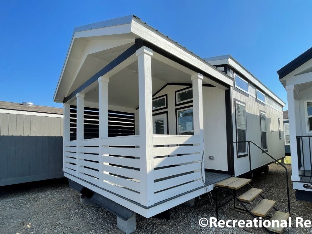Modern tiny home with white siding and black trim, featuring a cozy front porch with railings. Steps lead up to the entrance. Clear blue sky above.