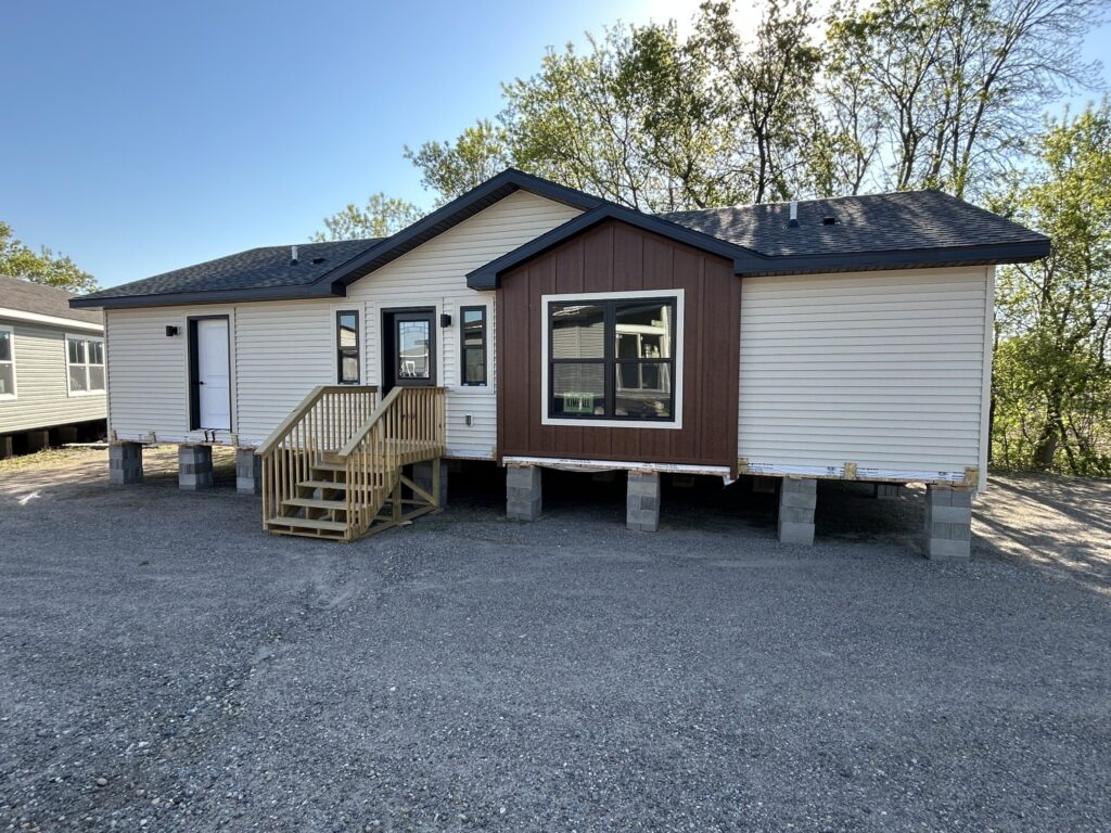 Modular home elevated on concrete blocks with white siding and brown accents. Front steps lead to the entrance. Trees in the background under a clear blue sky.