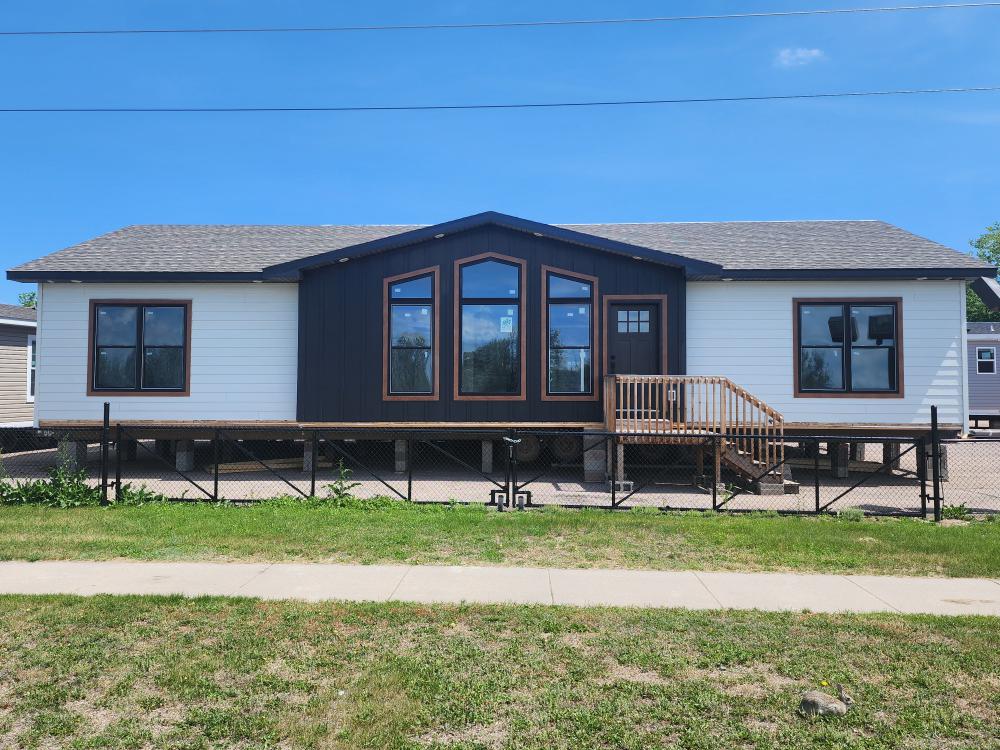 A modern, prefabricated house with a contrasting dark central facade, large windows, and a small porch. It is elevated slightly, with a chain-link fence and grassy lawn in front. Bright, clear sky in the background.