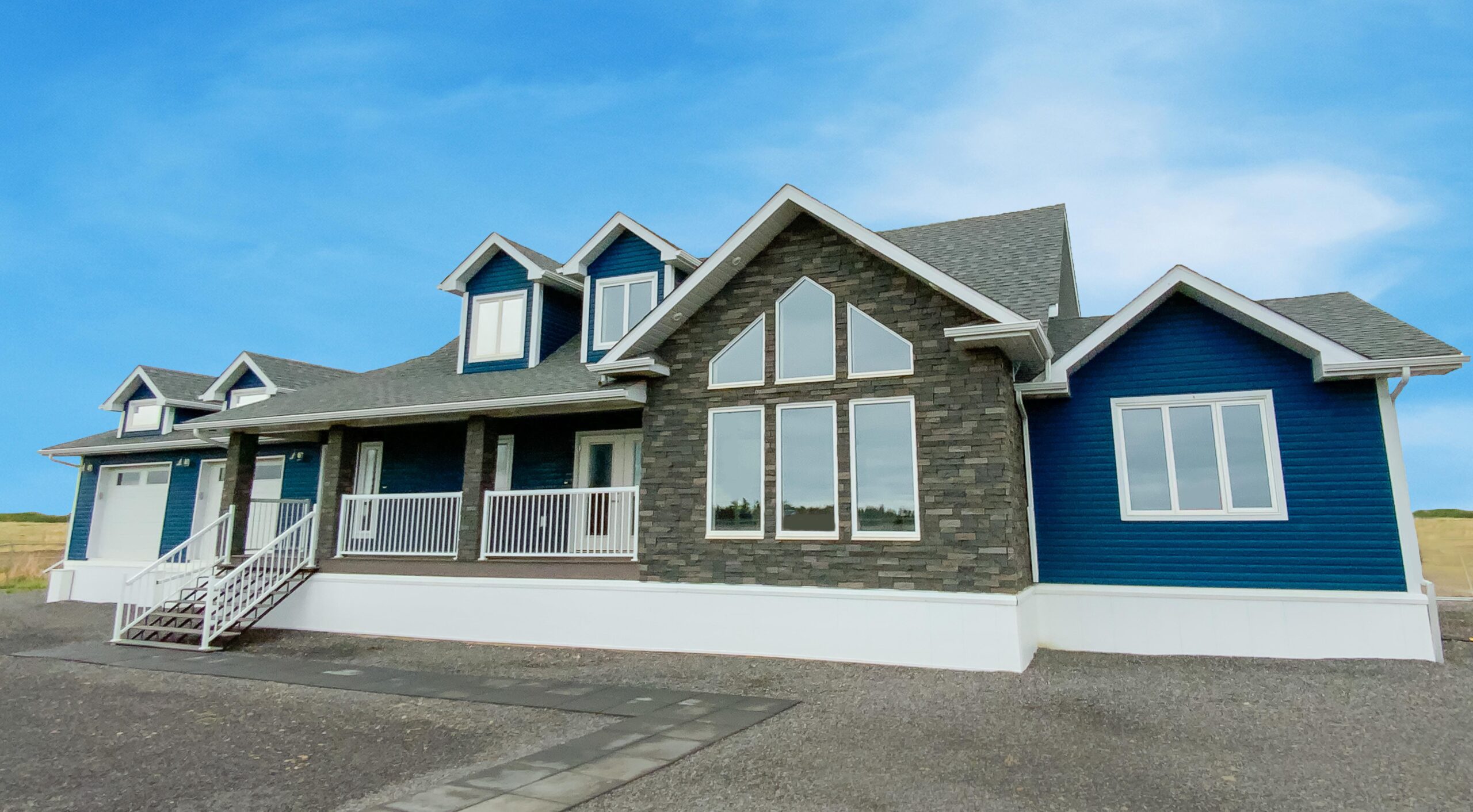 A modern blue and gray house with white trim, featuring large windows and a small porch with railings. The sky is clear and bright, conveying a serene ambiance.
