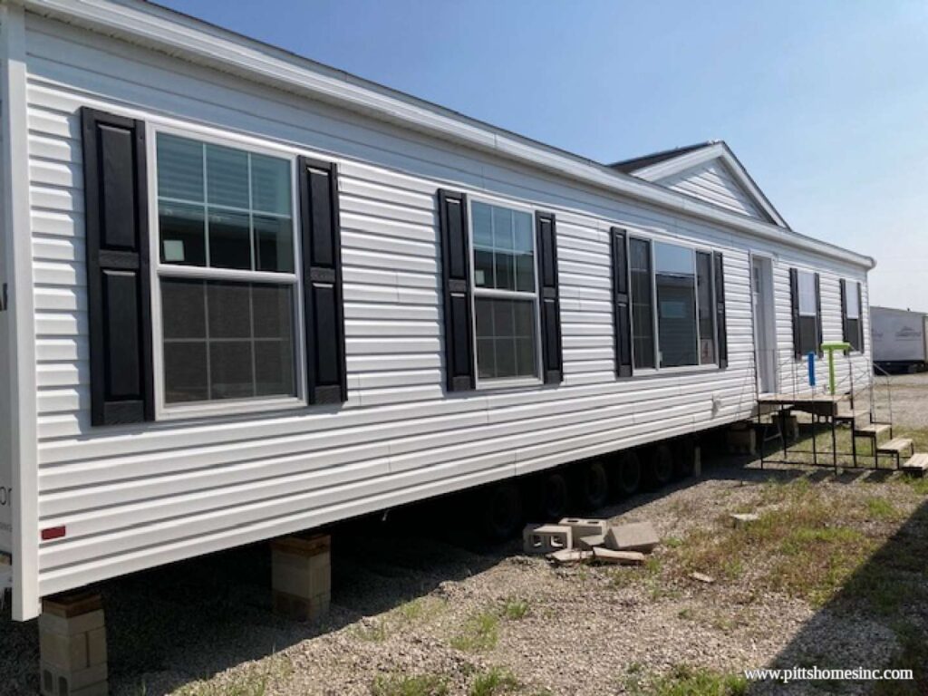White mobile home elevated on concrete blocks, six large windows with black shutters, and a small staircase leading to the door. Bright, sunny day.