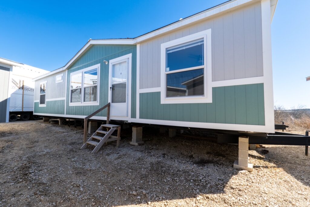 A green and gray manufactured home elevated on concrete blocks stands under a clear blue sky. Wooden steps lead to the front door, conveying a sense of simplicity.