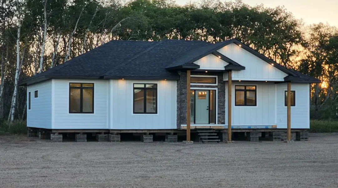 A modern, single-story house with a white facade and dark roof stands elevated on blocks. Warm lights illuminate the entrance, set against a backdrop of trees at sunset.