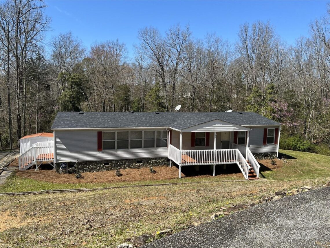 A single-story house with a black roof, white siding, and red shutters is surrounded by trees. It features a white porch and a circular deck on the left.