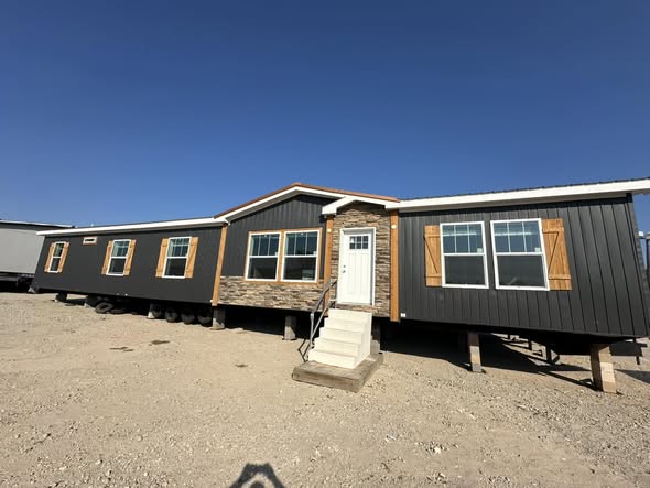 A large, dark-colored manufactured home with wood accents and stone veneer entry stands on a dirt lot under a clear blue sky. The mood is bright and open.