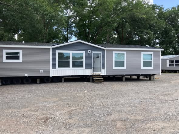 A gray mobile home with white trim sits on gravel, surrounded by lush green trees. Its front steps lead to a central door, creating a welcoming feel.