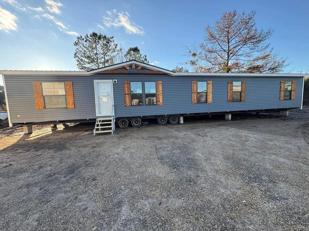 A large mobile home with blue siding and wooden shutters stands on wheels in a sunny, open area. Trees and a clear blue sky are in the background.