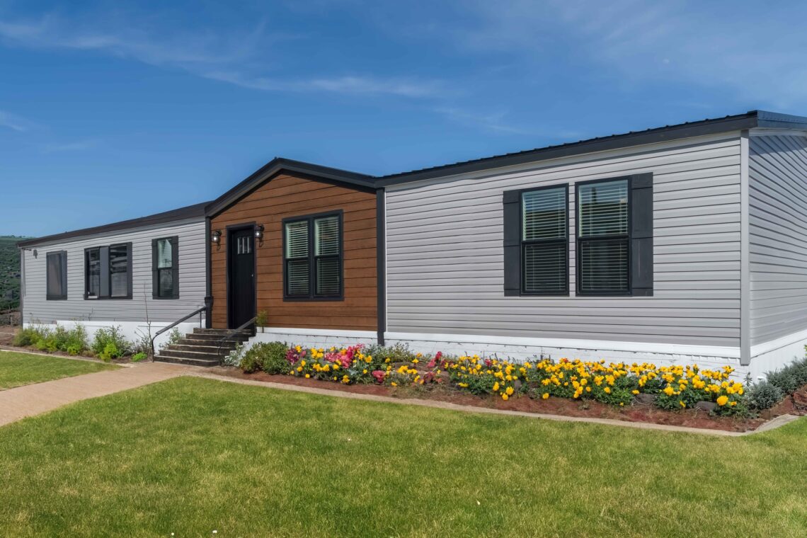 A modern, single-story house with gray and brown siding is surrounded by vibrant flowers and a lush green lawn under a clear blue sky.