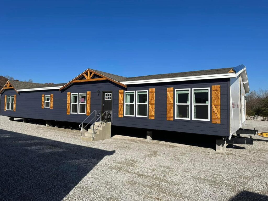 A long gray manufactured home with wood accents features a gabled roof and numerous windows. It sits on a gravel lot under a clear blue sky.