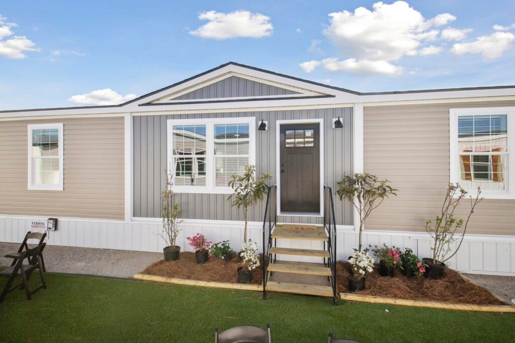 A beige and gray manufactured home with a central dark door, flanked by symmetrical windows. Plants and flowers line the entrance, creating a welcoming feel.
