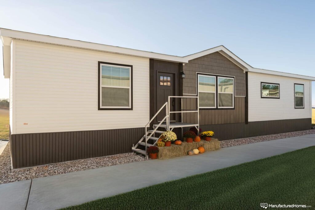 A modern manufactured home with a white exterior and dark trim sits under a clear sky. The porch is decorated with hay bales and colorful pumpkins.