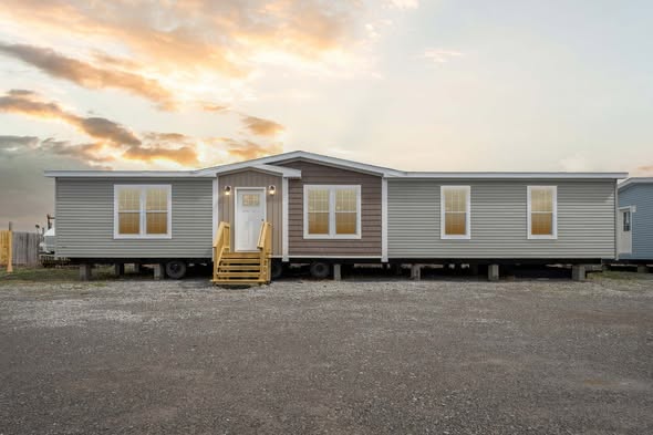 A modern, gray mobile home with a central white door and steps is set against a sunset sky. The gravel foreground enhances its simplicity and warmth.