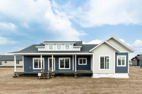Modern blue and white single-story house with a small porch, set on a dirt lot under a partly cloudy sky. The style is contemporary and inviting.
