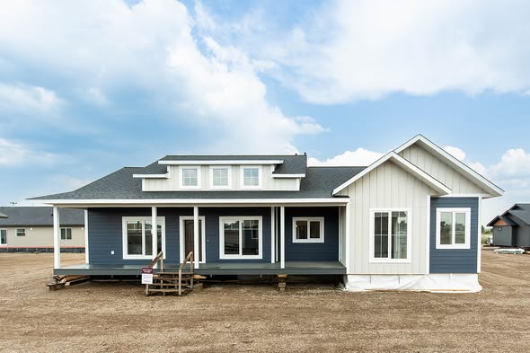 Modern blue and white single-story house with a small porch, set on a dirt lot under a partly cloudy sky. The style is contemporary and inviting.
