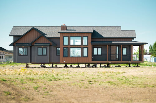 Modern brown and gray modular home elevated on blocks in an empty field under a clear blue sky, conveying a sense of new beginnings and construction.