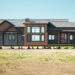 Modern brown and gray modular home elevated on blocks in an empty field under a clear blue sky, conveying a sense of new beginnings and construction.