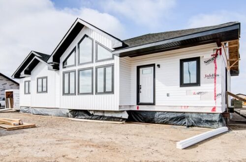 Single-story white house under construction, featuring large windows and black roof. Blue skies and scattered clouds create a hopeful, fresh atmosphere.