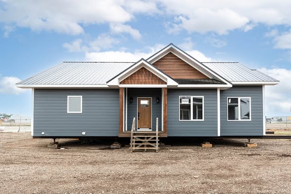 A newly constructed modular house with gray siding, white trim, and a metal roof. It has a central entrance with steps and large windows, set on a bare lot.
