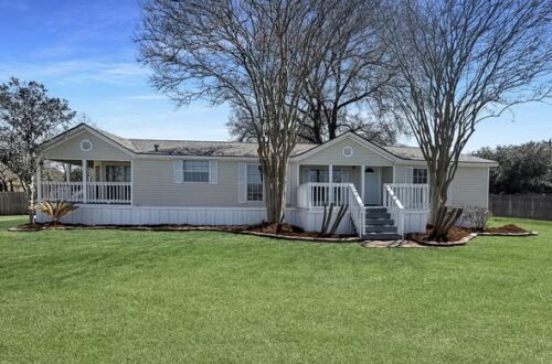 A charming single-story house with beige siding and white trim, set in a lush green lawn. Leafless trees frame the front porch. Clear blue sky above.