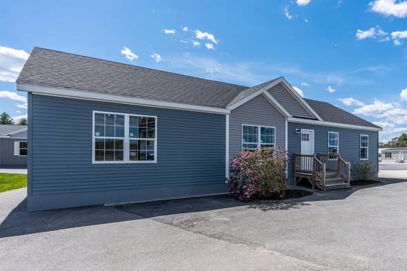 A single-story, blue-gray modular home under a clear blue sky. It has white trim, a gabled roof, front steps, and a small bush by the entrance.