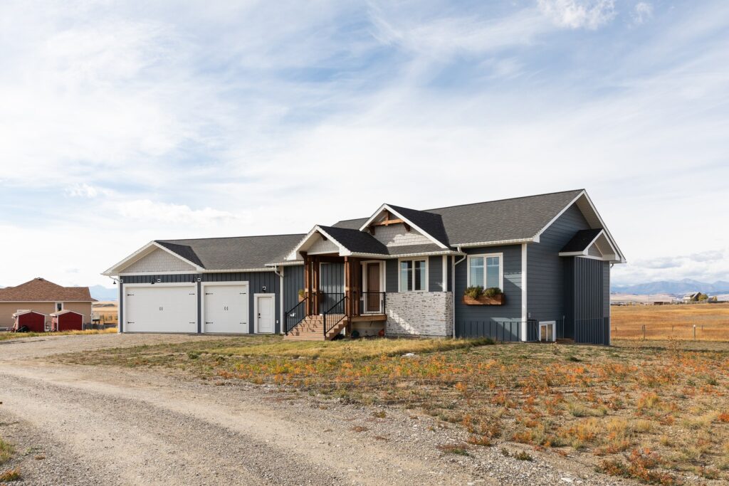 A modern grey house with a three-car garage on a gravel driveway, set in an open field under a wide blue sky. A serene, rural atmosphere.