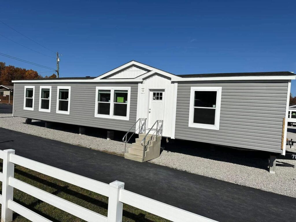 A modern gray mobile home with white trim sits on gravel, under a clear blue sky. A white fence lines the foreground, adding a suburban feel.