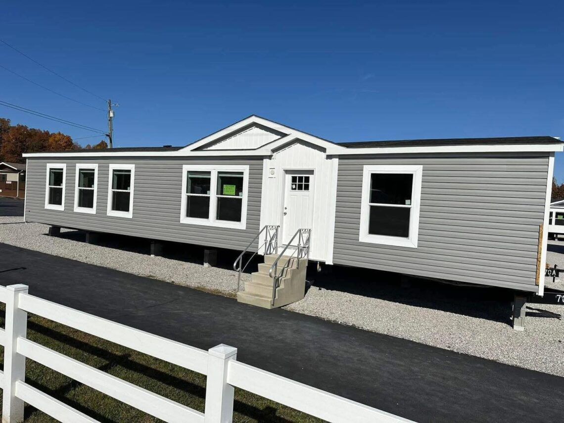 A modern gray mobile home with white trim sits on gravel, under a clear blue sky. A white fence lines the foreground, adding a suburban feel.