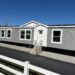 A modern gray mobile home with white trim sits on gravel, under a clear blue sky. A white fence lines the foreground, adding a suburban feel.