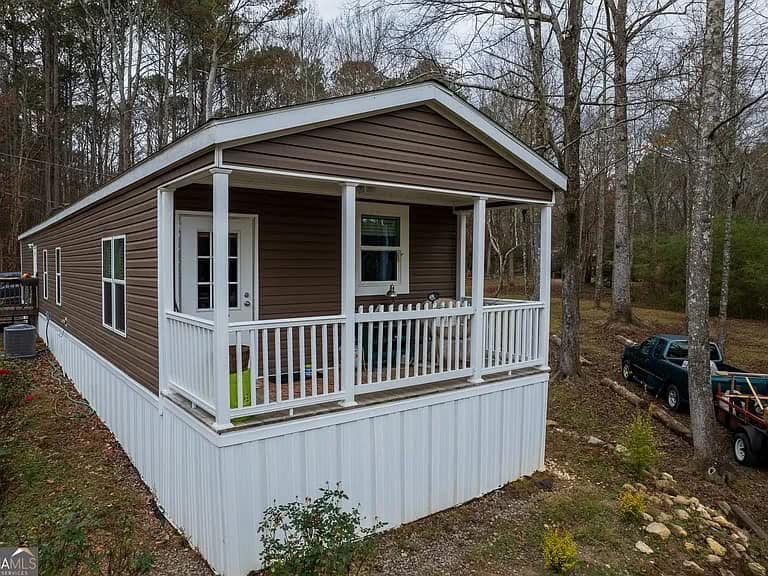 Brown mobile home with a white railing porch, nestled in a wooded area. Trucks are parked nearby, suggesting a rural, serene setting.