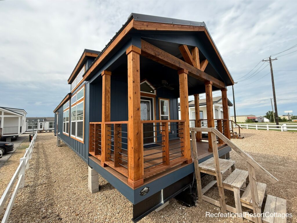 A modern tiny home with dark blue siding and warm wood accents. It features large windows and a small front porch with wooden railings, set on a gravel lot.