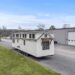 White tiny house on wheels with multiple windows, parked on a gravel driveway next to a large industrial building, under a clear blue sky.