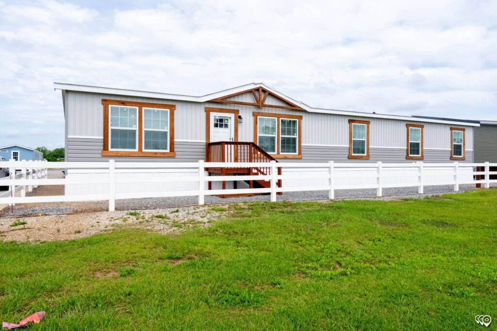 A long, gray and white mobile home with wooden trim, white railings, and steps is surrounded by a grassy yard. The sky is partly cloudy.