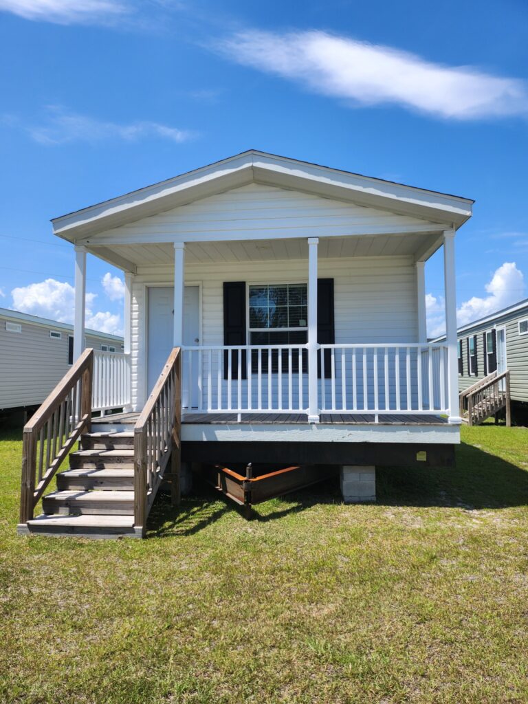 Small, white prefabricated home with a covered porch and steps, set on a grassy lot under a clear blue sky. The scene feels serene and welcoming.
