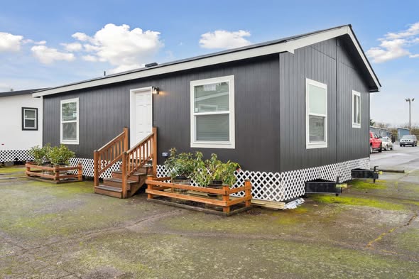Single-story prefabricated house with dark siding and white lattice trim. A small wooden porch leads to the front door, surrounded by potted plants.
