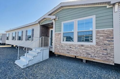 A modern manufactured home with a green and beige exterior features brick accents and multiple windows. A metal stairway leads to the entrance.