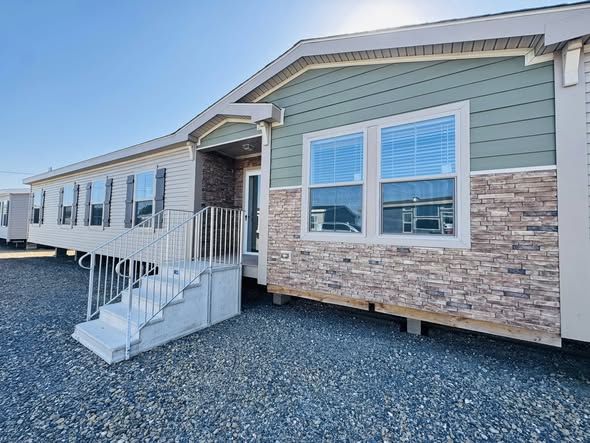 A modern manufactured home with a green and beige exterior features brick accents and multiple windows. A metal stairway leads to the entrance.