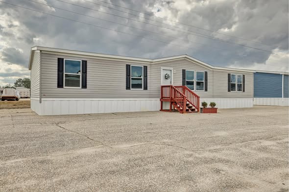 Single-story manufactured home with gray siding, white trim, and red steps. Overcast skies add a calm, neutral tone. Sparse landscape surrounds.