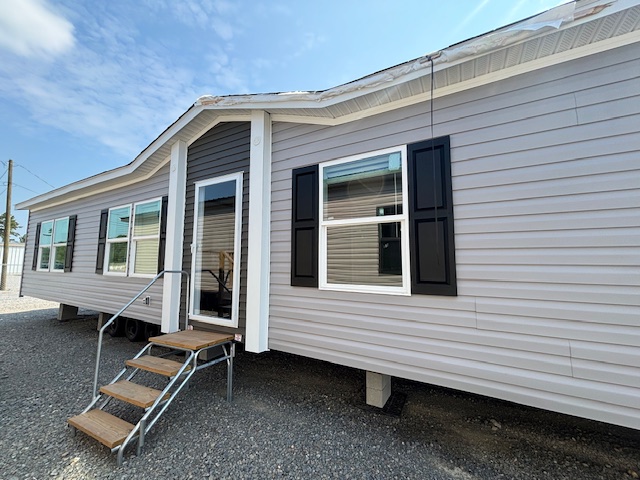 Modern mobile home with gray siding, black shutters, and a glass door, set on gravel with a small stairway. A clear blue sky above conveys calm.