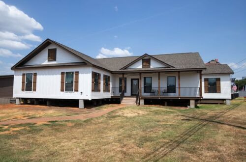 Large, elevated white house with a brown roof, front porch, and wooden shutters set against a blue sky. Surrounding lawn is dry and patchy.