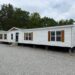 A large, white mobile home with wooden shutters and multiple windows is set on a gravel lot. Surrounded by trees, the scene feels serene and rural.
