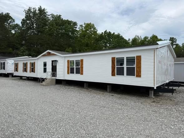 A large, white mobile home with wooden shutters and multiple windows is set on a gravel lot. Surrounded by trees, the scene feels serene and rural.